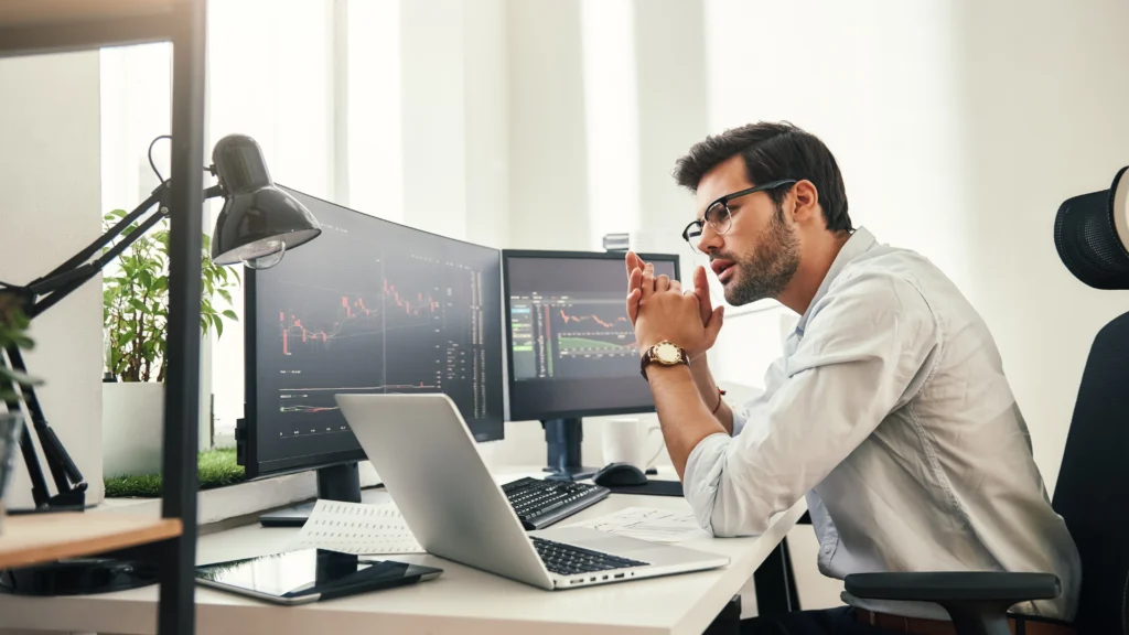 13d660b4-08fb-435a-bdf1-fe7582531459 – PU Prime | More Than Trading man looking at laptop and computer monitors with cfd trading charts on them, while sitting down
