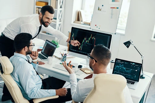 dd73158c6a044331805647935c7fb903 – PU Prime | More Than Trading three businessmen engaging in a discussion regarding trading indices shown on the monitor screen, and two laptops